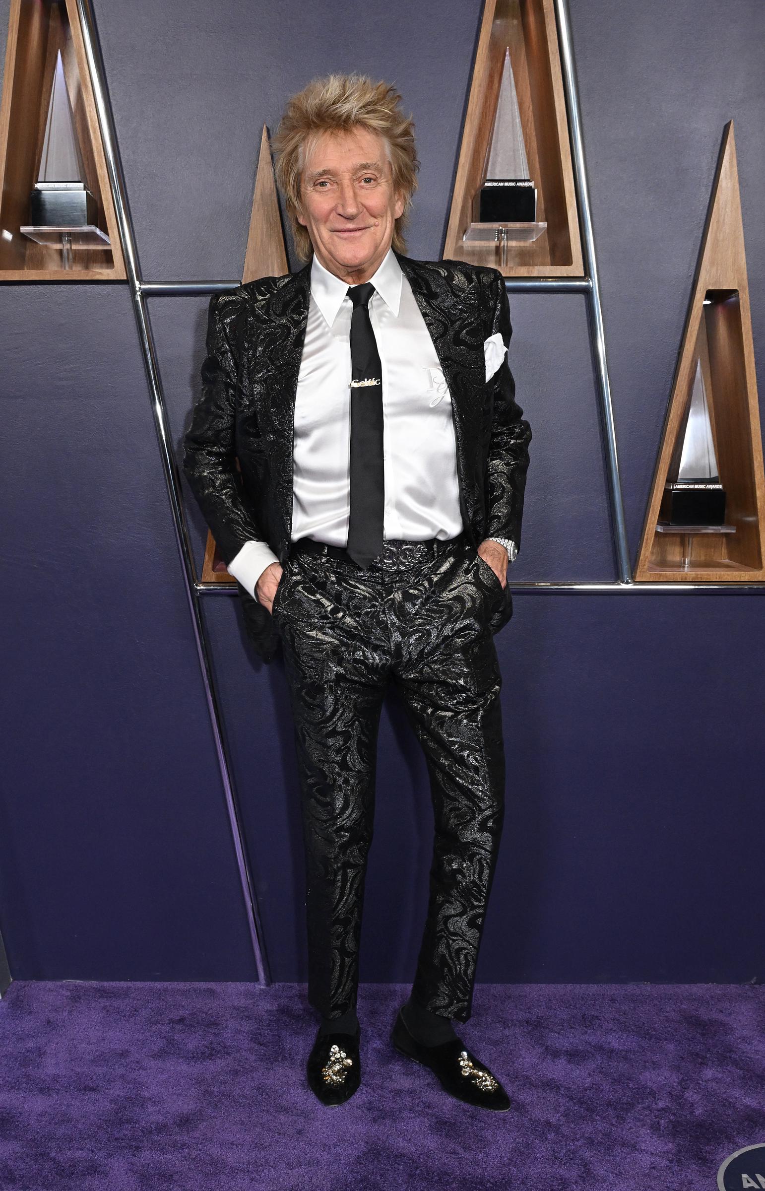 Rod Stewart posed coolly on the purple carpet in a metallic black brocade suit. Matching pants and embroidered velvet loafers enhanced the polished look, while a bold belt buckle added a retro-glam finish to the timeless ensemble.