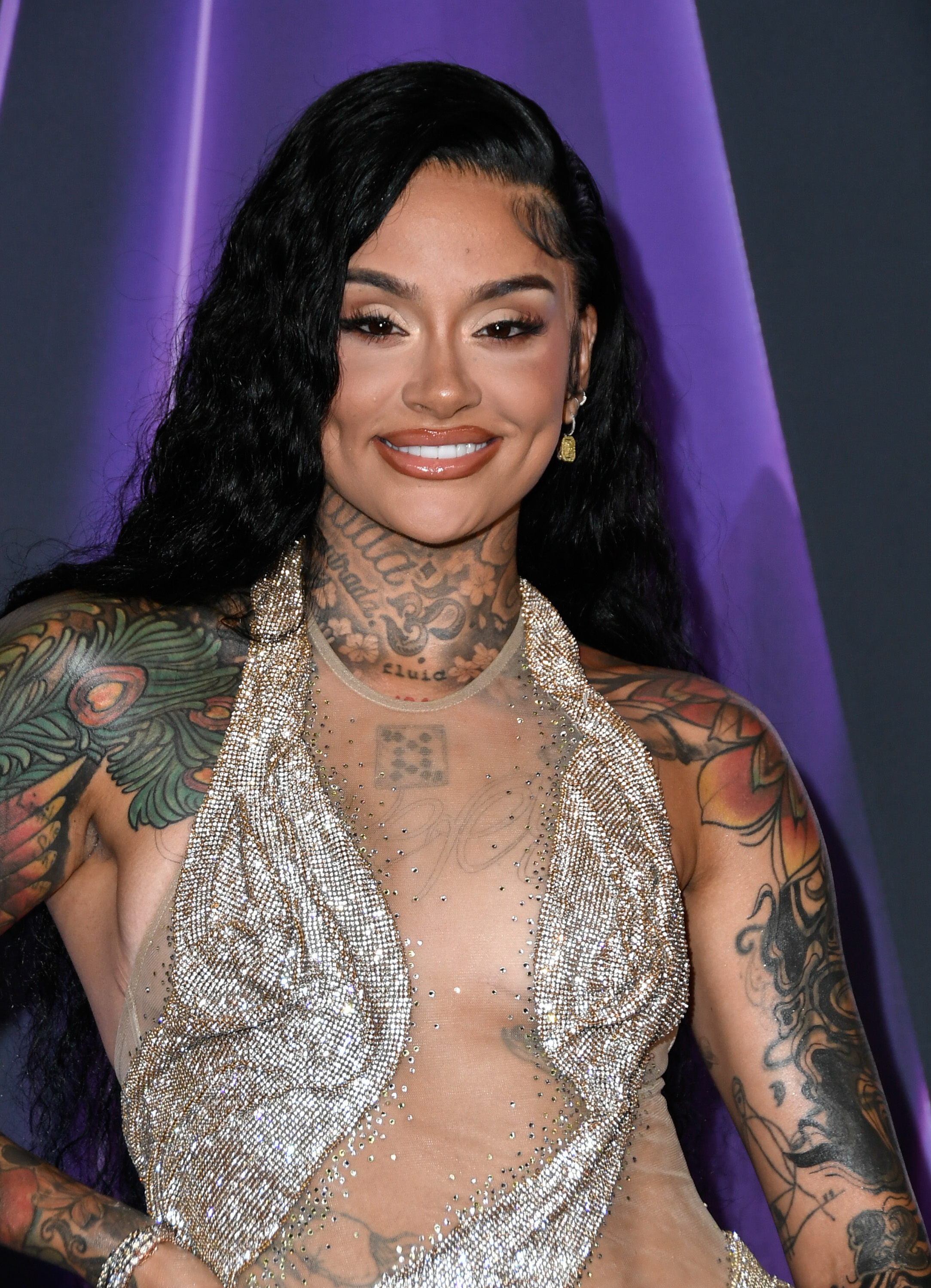 Flashing a radiant smile, Kehlani stunned at the 2025 AMAs in a sheer, mesh-based gown overlaid with glimmering silver draping that swirled across her chest. A sparkling neckline framed her tattoos, blending sensuality with self-expression.