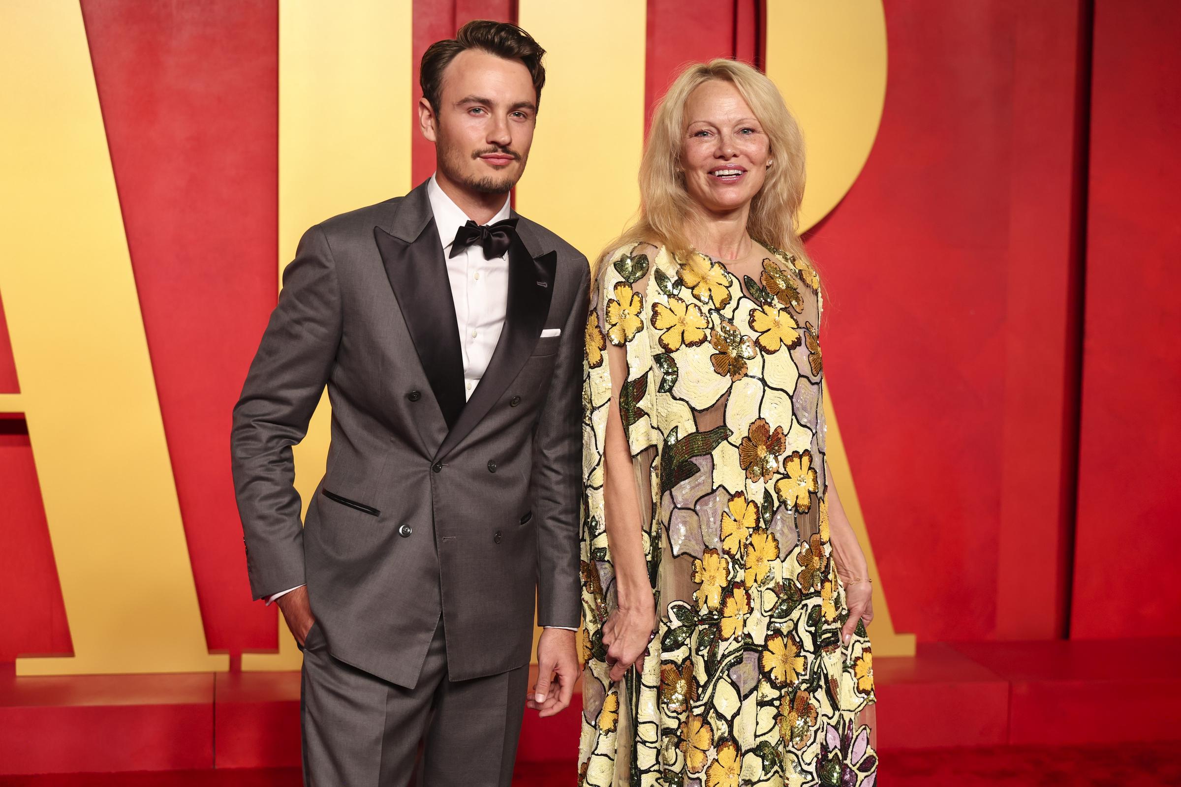 Brandon Thomas Lee and Pamela Anderson attend the Vanity Fair Oscar Party 2024. Lee wears a grey tuxedo with black satin lapels and a bow tie, while Anderson appears in a floral sequin-embellished dress with yellow and white tones.