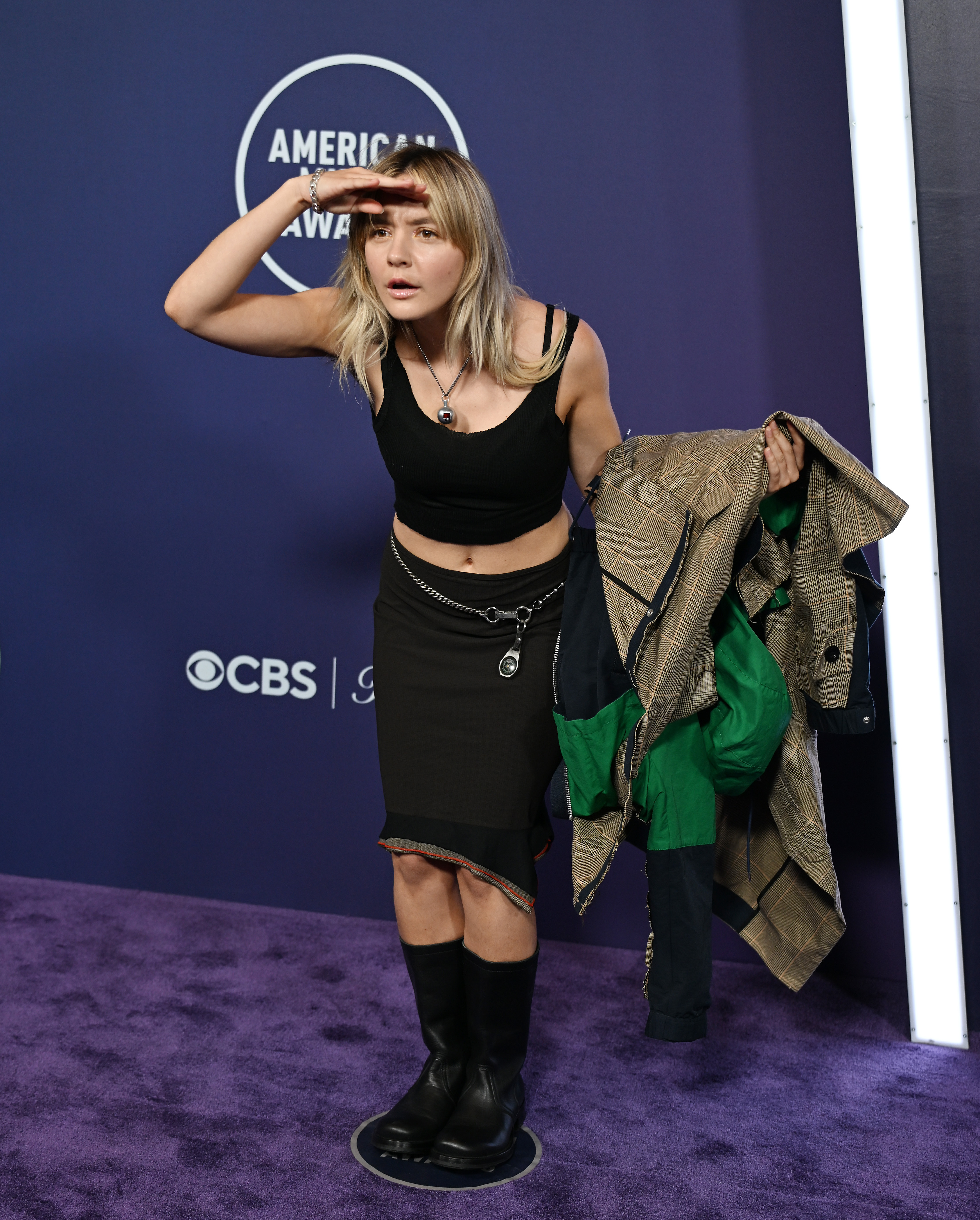 At the 2025 AMAs, Dora Jar revealed a black cropped tank and asymmetrical midi skirt beneath her layers. The look was styled with mid-calf boots, silver hardware, and a looped keychain belt, adding structure to the minimalist silhouette.