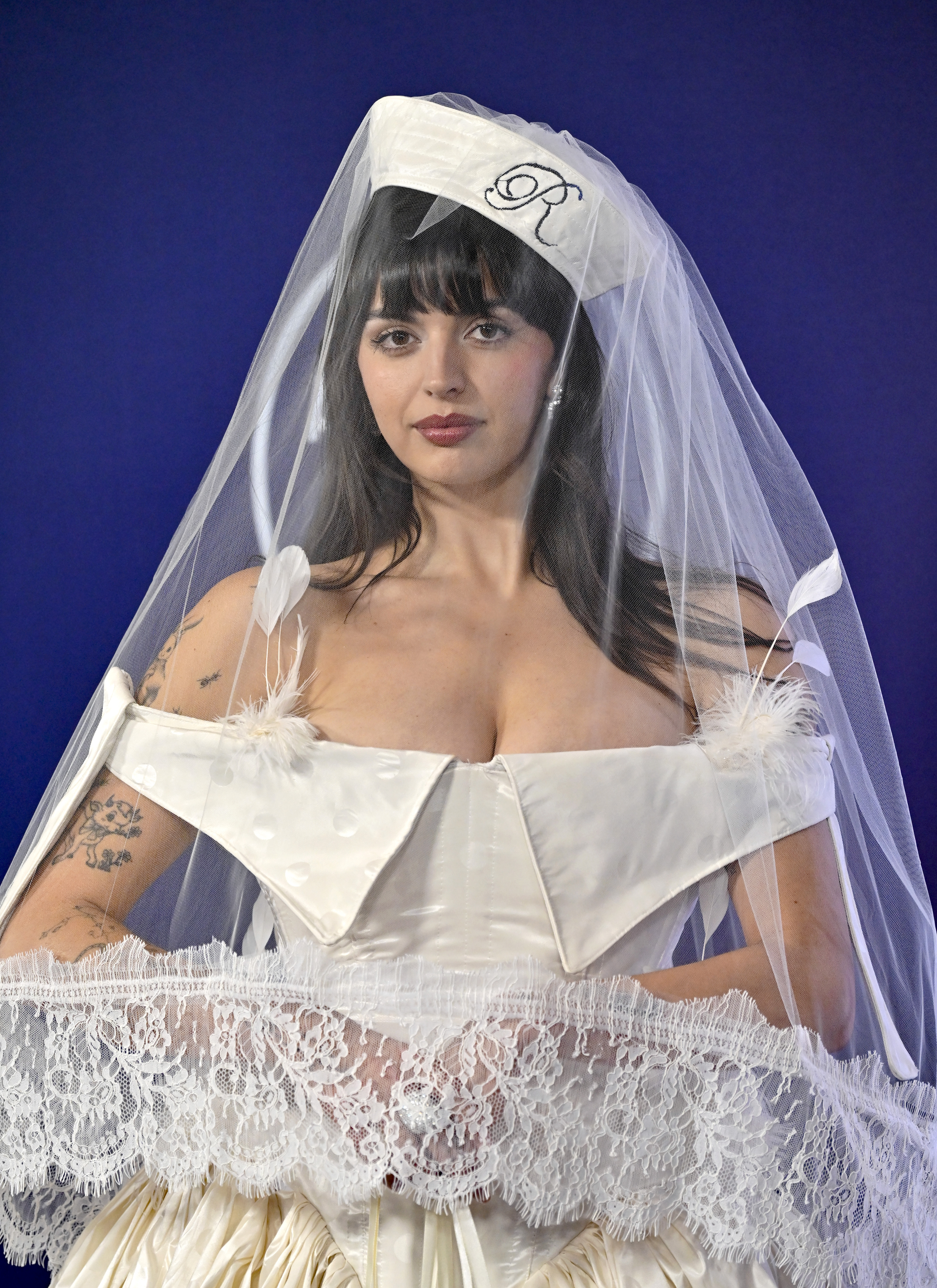 Beneath her embroidered beret and tulle veil, Rebecca Black wore an off-the-shoulder dress trimmed with delicate feathers. A flared, tea-length skirt completed the playful, vintage-glam look — bridal, but with a bold twist.