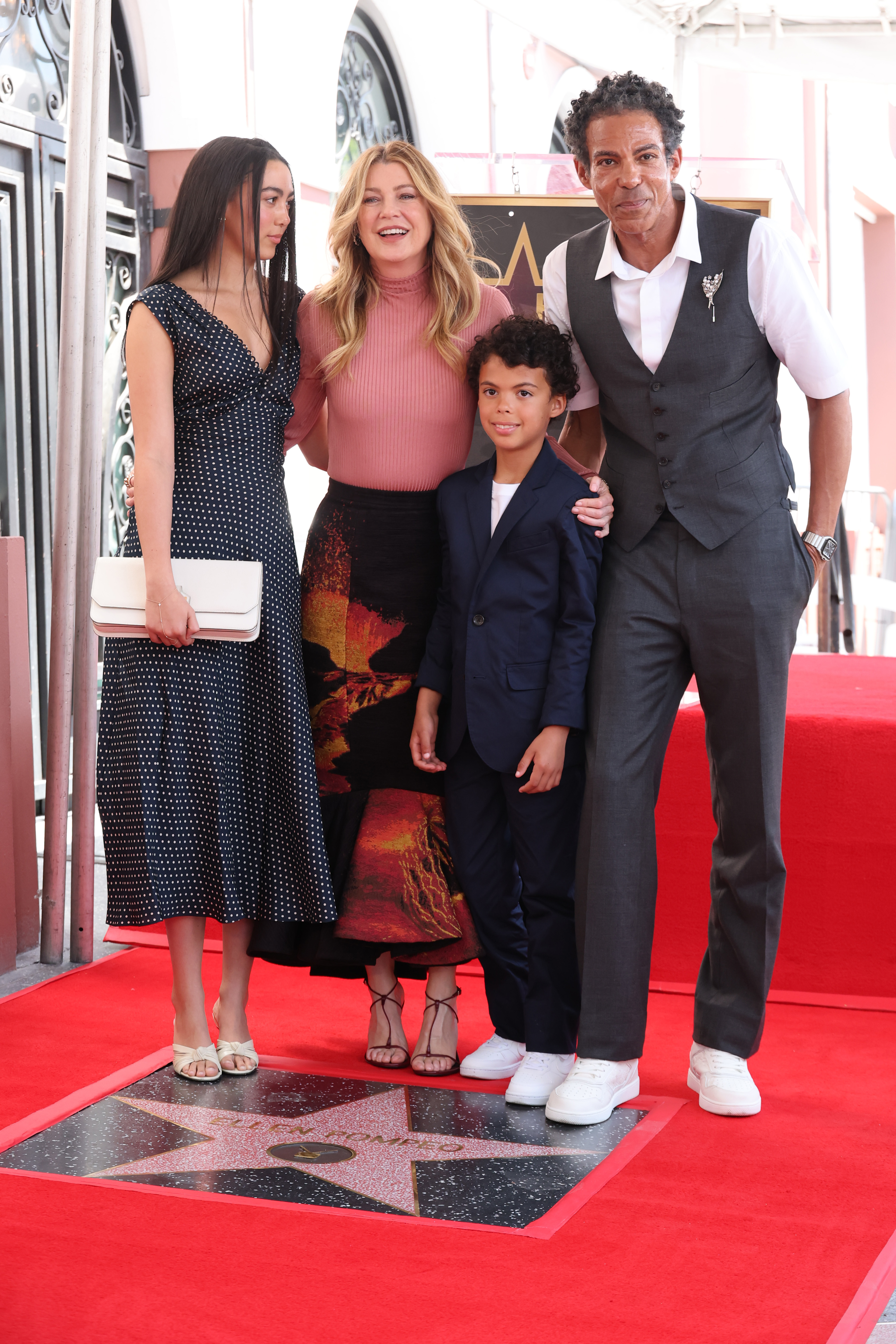 The family stands proudly over Ellen’s star, all sharing a joyful moment. Ellen beams with a wide smile, while Chris looks on proudly with a calm, confident expression. Their daughter gazes at Ellen with quiet affection, and their son offers a sweet, shy smile to the camera.