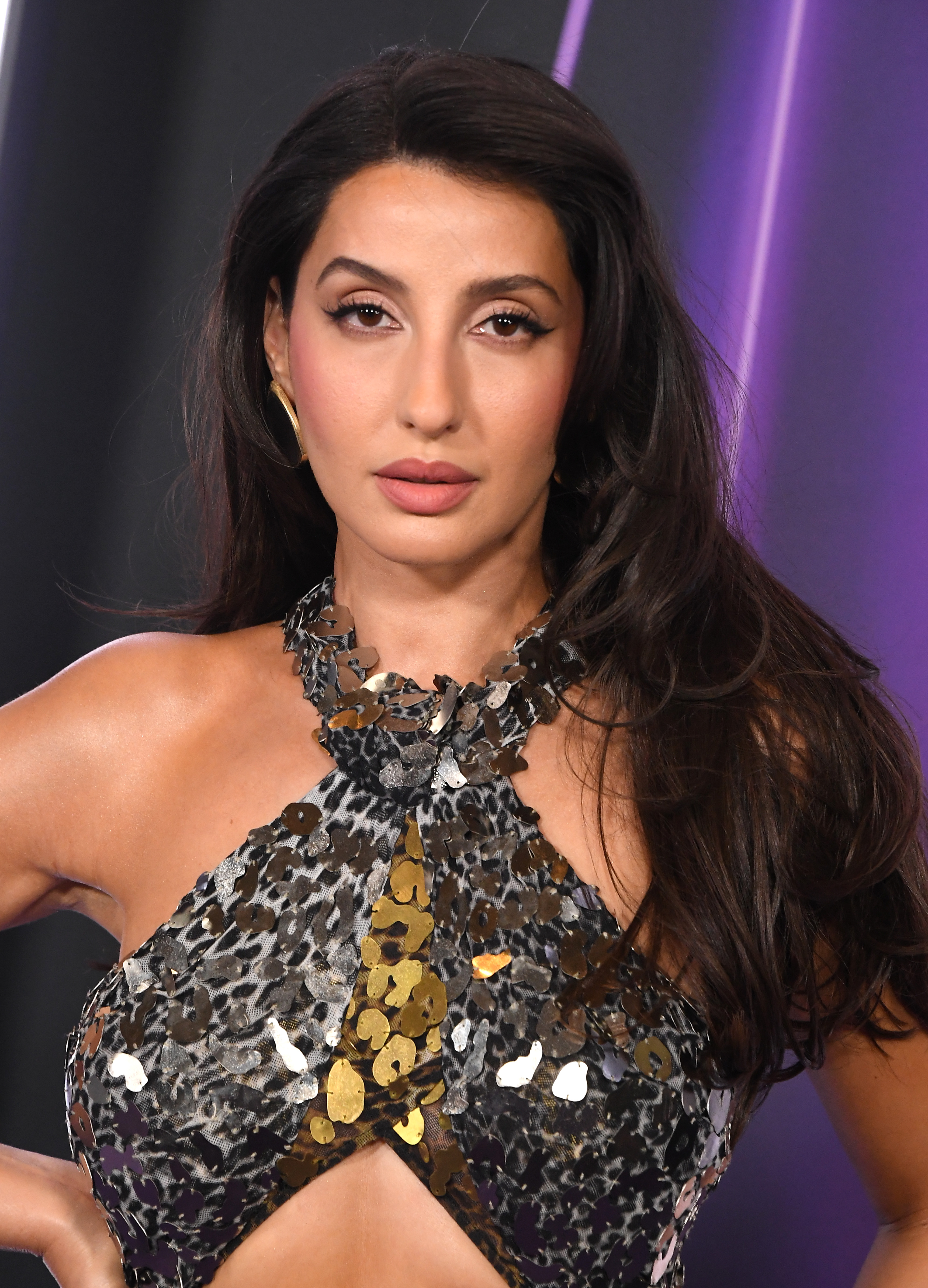 Nora Fatehi at the 2025 AMAs wore a halter-style gown with metallic paillettes in mixed tones. Her makeup featured defined eyes and a nude lip, paired with softly styled waves and gold hoop earrings.